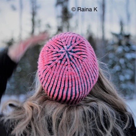 White woman looking away from the camera with long blonde hair. She wears a pink knitted hat and there are out of focus snowy trees in the background.