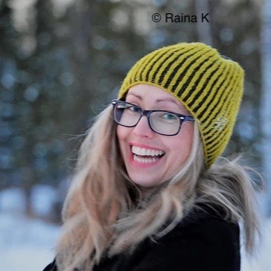 White woman looking directly into camera and laughing with long blonde hair and black glasses. She wears a green/yellow knitted hat and there are out of focus snowy trees in the background.