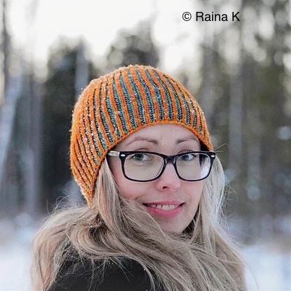 White woman looking directly into camera with long blonde hair and black glasses. She wears an orange knitted hat and there are out of focus snowy trees in the background.